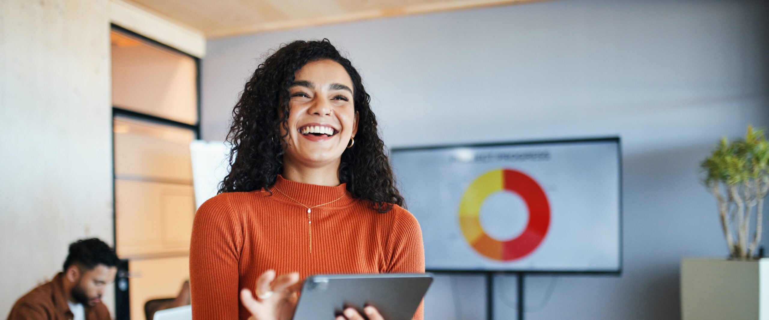 Smiling businesswoman with curly hair reads talent acquisition data on a tablet. A pie chart is displayed on a screen behind her. A colleague is working in the background, creating a collaborative office environment.