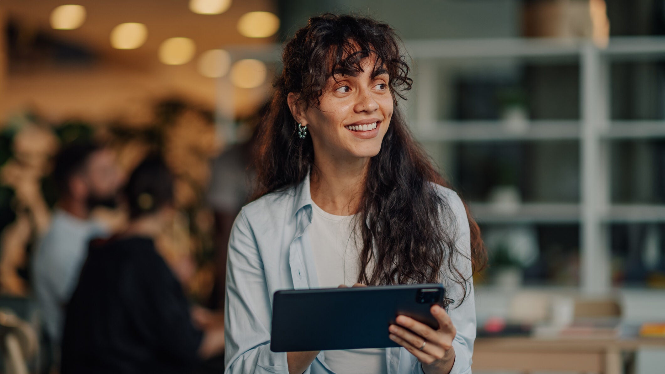 Portrait of a young businesswoman using a digital tablet, smiling and looking away in a modern office with colleagues in the background
