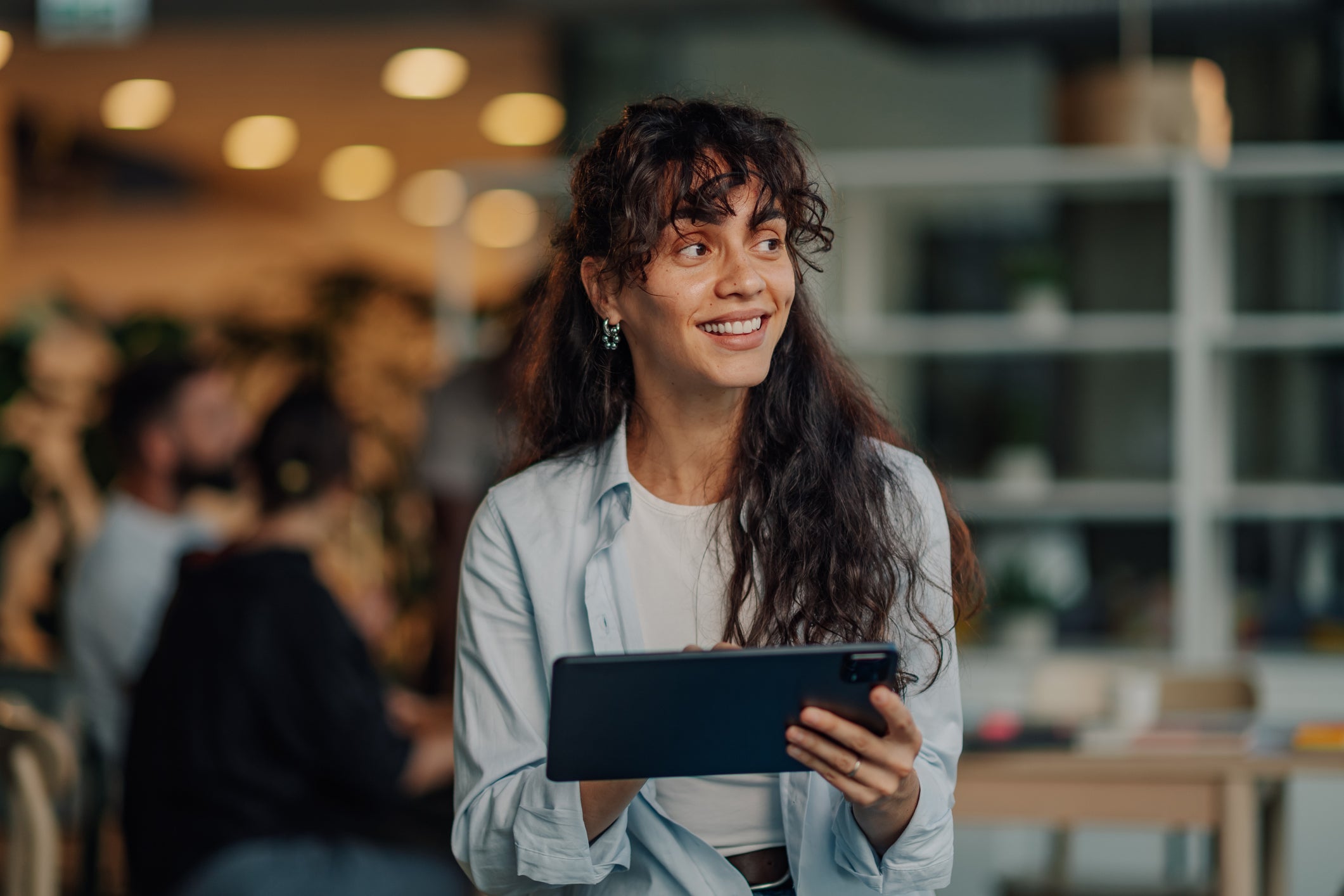 Portrait of a young businesswoman using a digital tablet, smiling and looking away in a modern office with colleagues in the background