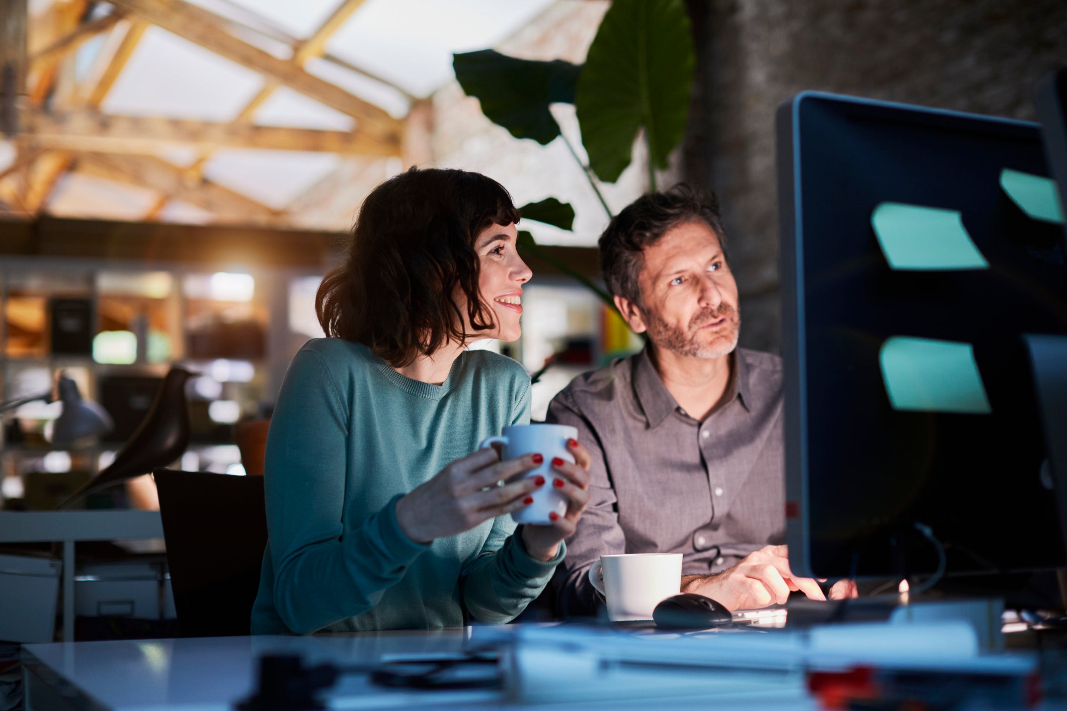 A male and a female colleague drinking coffee, looking at their computer screen, and smiling in their office.